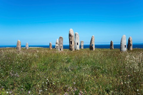 Group of standing stones in a meadow, surrounded by nature and under a blue sky with the sea in the