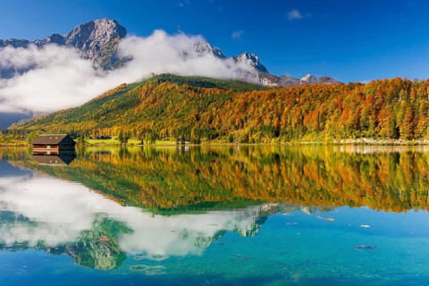 Lake Almsee in autumn with a forest reflected in the lake. A slowly dissipating fog moves over the