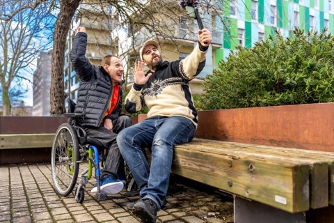 Young man in a wheelchair and his videographer friend are recording a video blog in a city park,