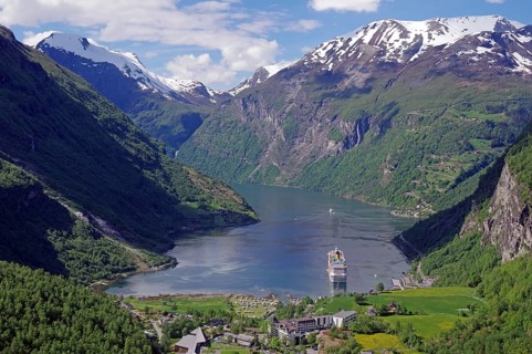 A fjord with a cruise ship surrounded by green mountains with snow-capped peaks under a blue sky