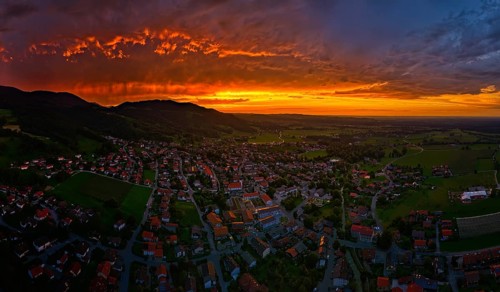 A village under a dramatic sunset sky with glowing clouds and mountains in the background, Bad