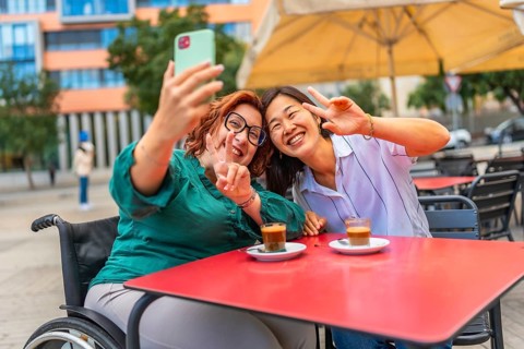 Cheerful disabled woman and asian female friend gesturing success taking selfie sitting in an