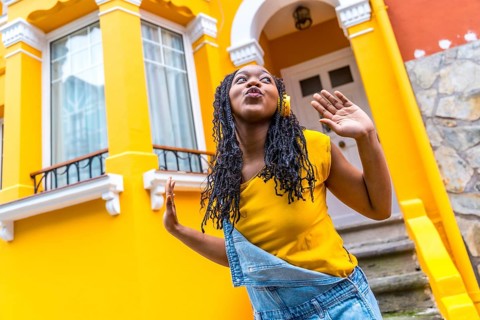 Cute african woman dancing listening to music with headphones next to a yellow house outdoors