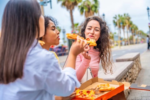 Three female diverse tourists chatting and sharing pizza sitting in the street