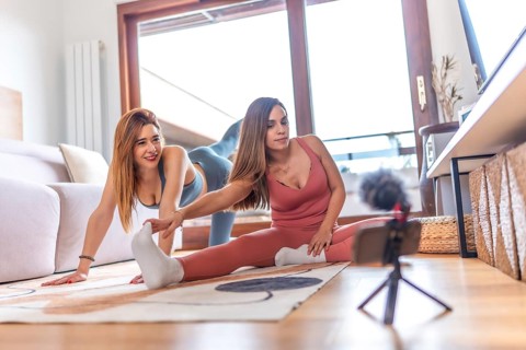 Two young women recording an online yoga class at home, performing stretching exercises in the