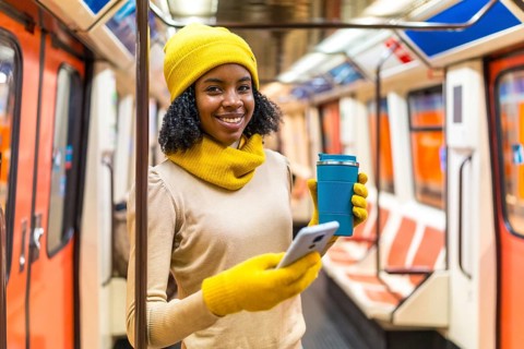 Smiling woman wearing yellow winter clothes using smartphone and drinking coffee inside subway