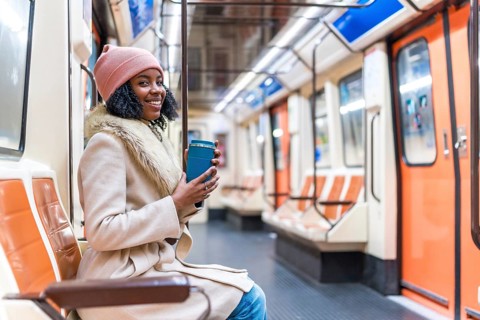 Smiling young black woman wearing winter clothes holding reusable coffee cup while riding subway