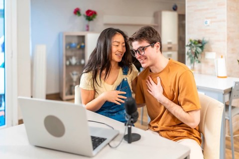Young multiethnic couple sitting together at home, using a laptop and microphone to record their