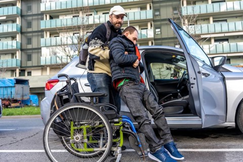 Caregiver lifting a disabled young man from his wheelchair and helping him to get into a car parked