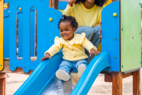 Cute african girl sliding in slide in a colorful playground next to her mother