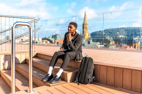 Pensive african young male traveler writing notes sitting on a city viewpoint during sunny day