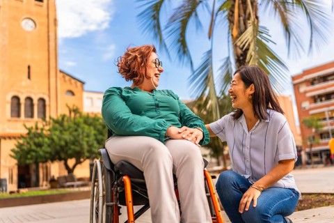 Asian caregiver holding hand with disabled woman in wheelchair outdoors in a beauty city square