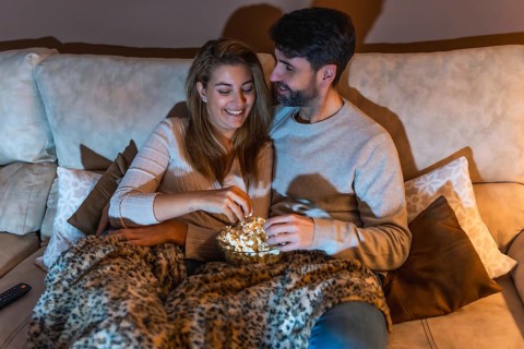 Romantic couple sharing a bowl of popcorn while watching a movie on the sofa at night, enjoying a