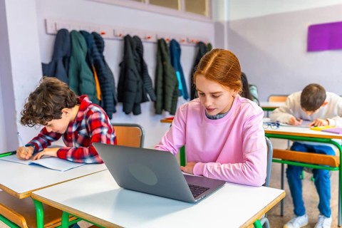Elementary school students studying at individual desks, girl using laptop, boy writing in