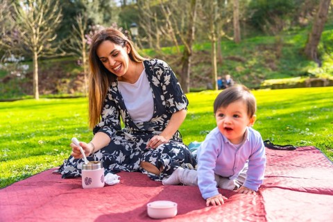 Happy mother preparing a meal from a thermos for her baby during a picnic on a blanket in a sunny