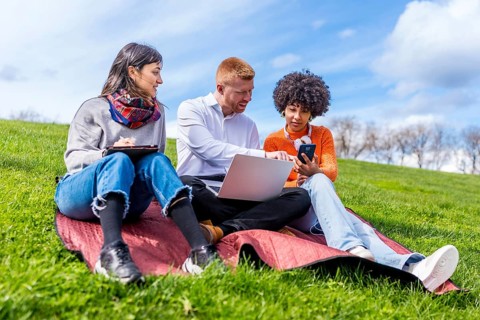 Three students collaborating on a project, using laptop, tablet, and smartphone, enjoying sunny