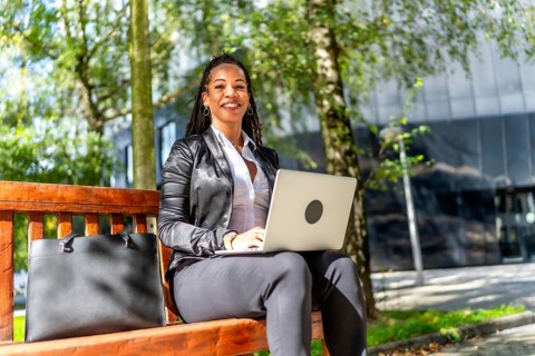 Latin businesswoman smiling at camera while sitting on city bench working with laptop
