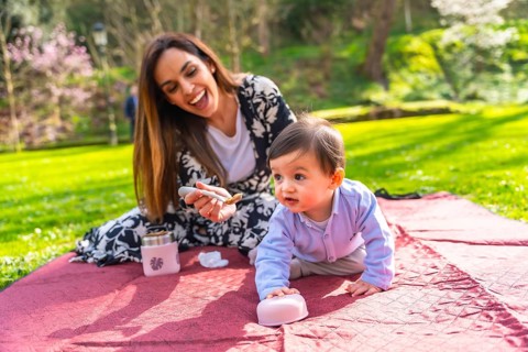 Happy mother feeding her baby solid food with a spoon, sitting on a blanket during a picnic in a