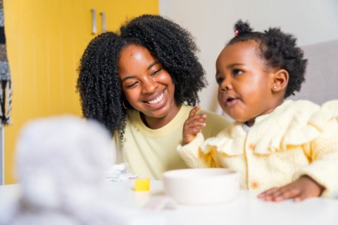 African toddler eating in the living room next to her smiling mother at home