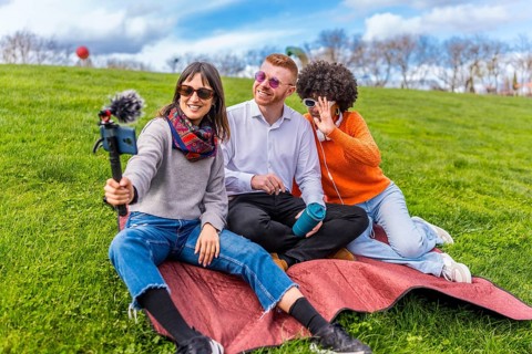Three content creators are sitting on blanket in park, filming video blog using smartphone with