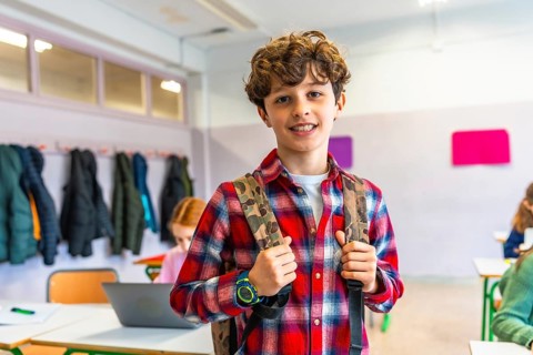 Portrait of a smiling preteen student wearing backpack in a classroom with classmates studying on