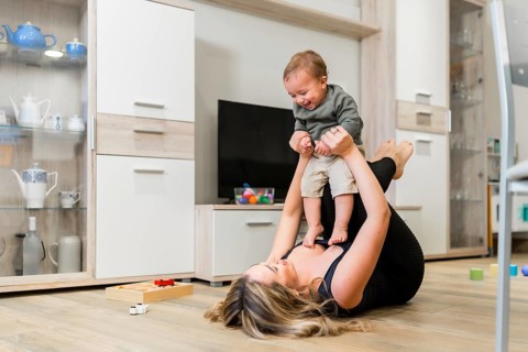 Young mother lying on the floor lifting her baby in the air while playing together in the living