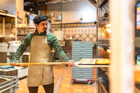 Latin male baker using wooden peel to remove baked pastries from a traditional oven