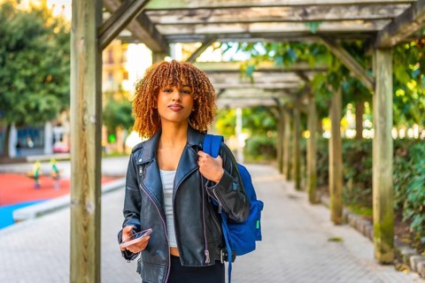 Happy latin female student walking along the city carrying bag and using smartphone