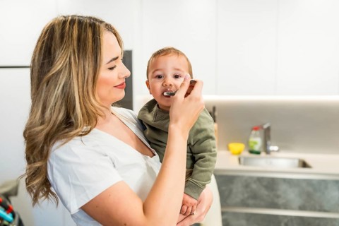 Loving mother feeding her baby son with a spoon in a bright modern kitchen, enjoying a tender
