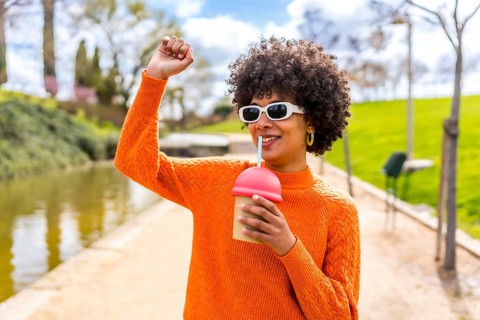 Happy young woman with curly hair and orange sweater, enjoying a smoothie in a park by the river,