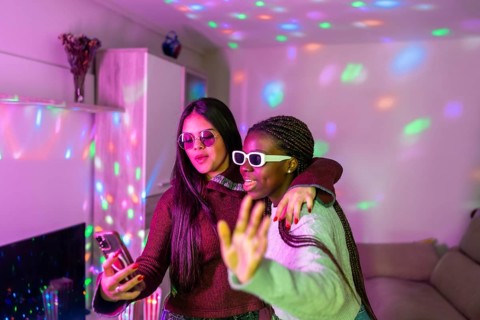 Two teenage girls wearing sunglasses are embracing and taking a selfie with smartphone at a home