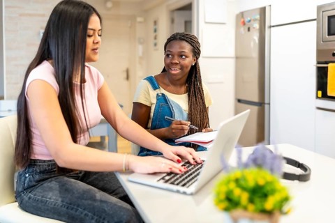 Two teenage girls collaborating on homework, using a laptop and taking notes in a modern kitchen