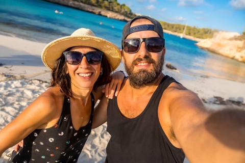 A man and woman are posing for a picture on a beach. The man is wearing a black tank top and