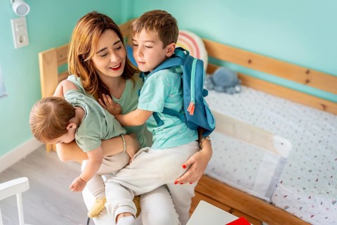 Mother embracing and holding a school boy and baby sitting together in the bedroom before going to