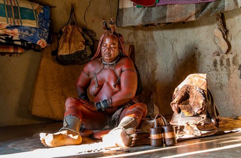 Traditional Himba woman applying ochre paint for body colouring, interior of the first woman's mud