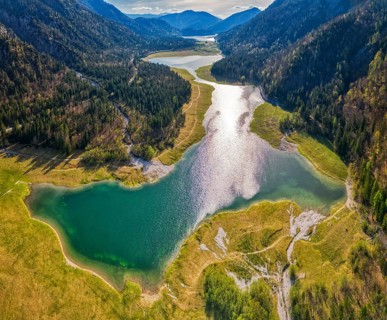 Green mountain landscape with river and lake, sun reflection in the water, surrounded by forests,