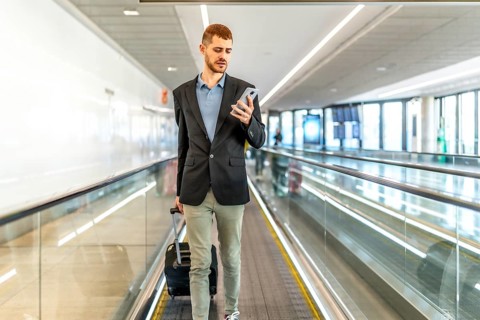 Businessman pulling suitcase and using smartphone while walking on moving walkway at airport