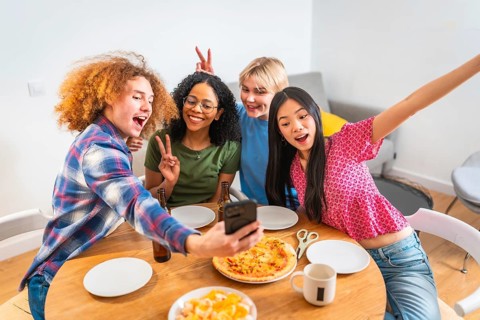 Four diverse friends capture a fun moment with a selfie while having pizza, snacks, and drinks at a