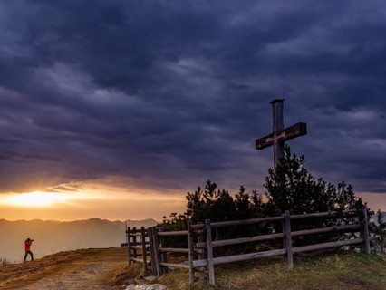 Photographer next to the summit cross on the Roßfeld in a dramatically overcast sky, the sun shines