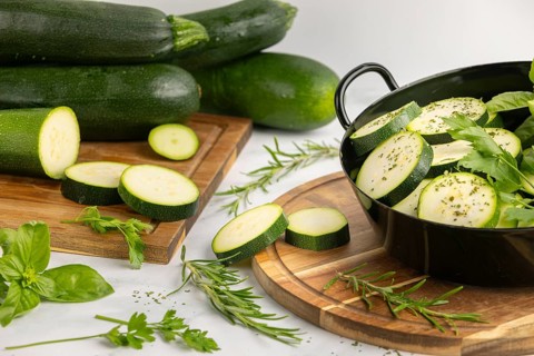 Sliced and seasoned courgettes with fresh herbs in a black pot, next to stacked courgettes on a