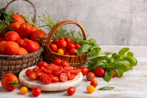 Sliced tomatoes on a white marble slab, surrounded by fresh tomatoes in baskets and herbs