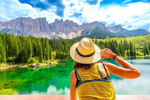 Woman wearing straw hat admiring lake carezza with latemar mountain range in background, dolomites,