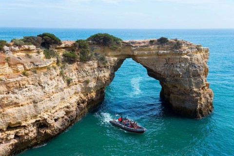 A boat with tourists sails through a natural rock arch over the clear blue sea, Arco de Albandeira,
