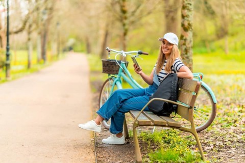 Happy tourist is using her smartphone while resting on a bench in a park with her bicycle next to