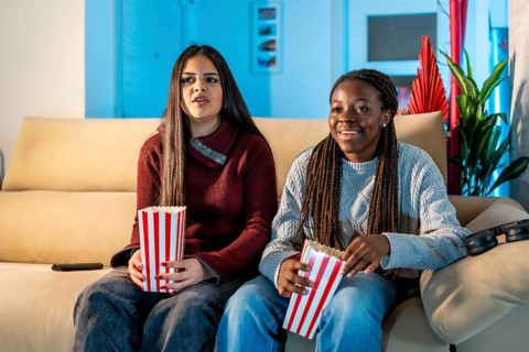 Two teenage girls enjoying a movie night at home, sitting on a comfortable sofa, eating popcorn