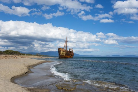 An abandoned, rusty shipwreck lies on a sunny beach with blue sky and clouds, Shipwreck Dimitrios,