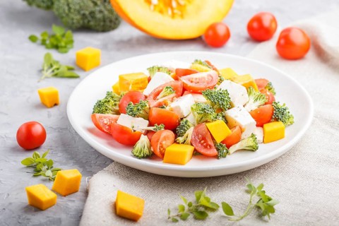 Vegetarian salad with broccoli, tomatoes, feta cheese, and pumpkin on white ceramic plate on a gray