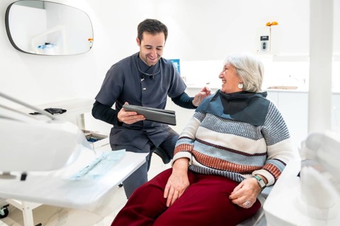 Dentist showing something on tablet to smiling senior woman sitting in dentist chair, during