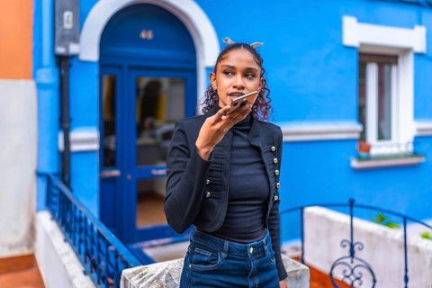 African american woman sending a voice message with smartphone next to colorful blue house