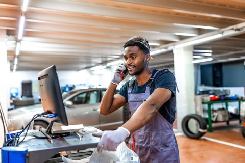 Young mechanic wearing gray uniform and protective glasses using computer and talking on phone